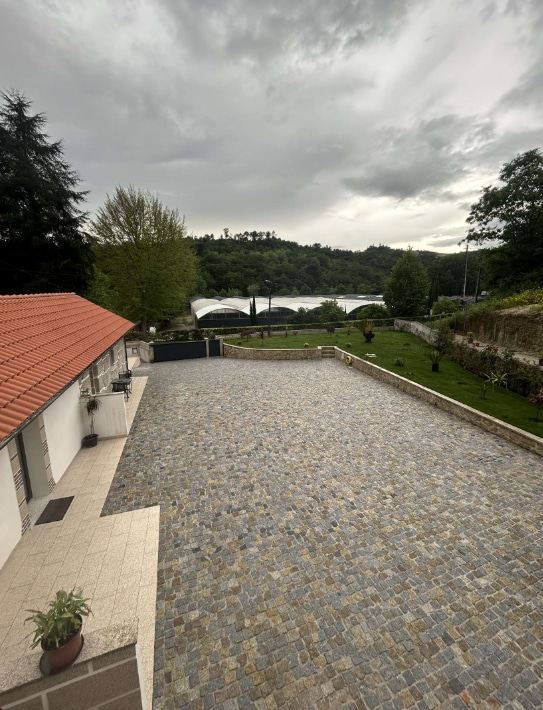 A spacious outdoor area is presented with a cobblestone surface, bordered by greenery and trees. The view features hills in the background, visible structures, and a serene atmosphere under a cloudy sky.