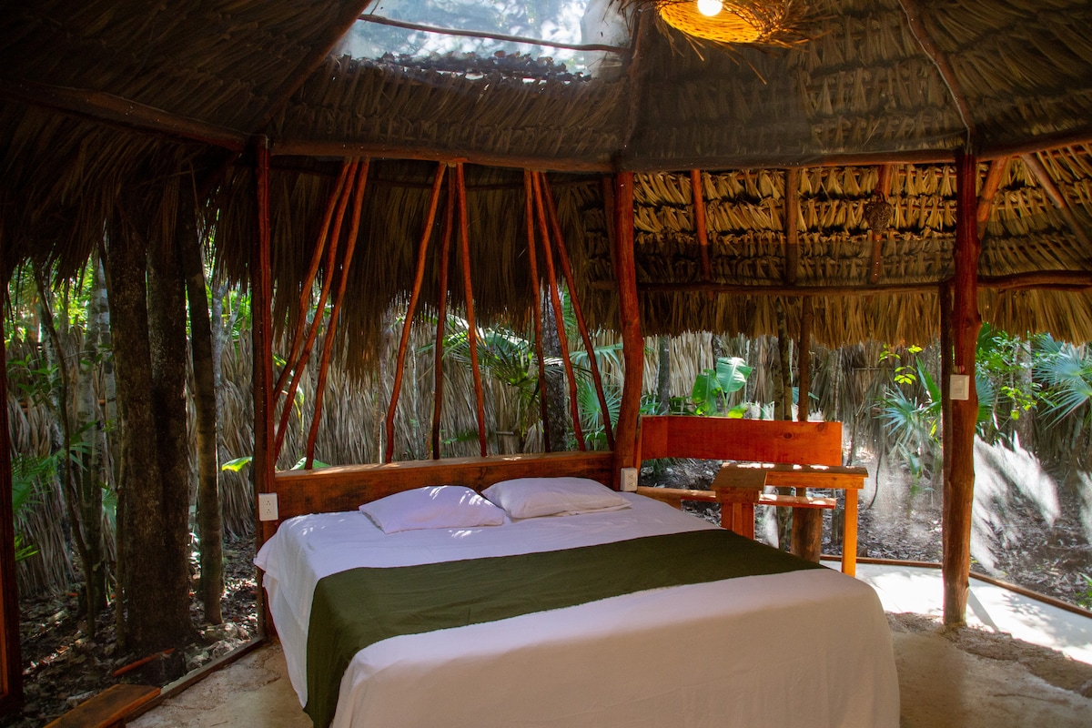 A spacious sleeping area is displayed with a king-size bed, dressed in crisp white linens and a dark green throw. The natural wood structure features a thatched roof and open walls, allowing sunlight and fresh air to flow through, surrounded by lush greenery.