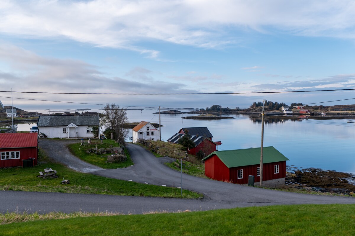 A serene coastal landscape features a mix of traditional red and white houses surrounding a calm waterway. The scene is framed by gentle hills and a cloud-filled sky reflecting on the water's surface. Power lines stretch across the tranquil atmosphere, enhancing the rural charm.