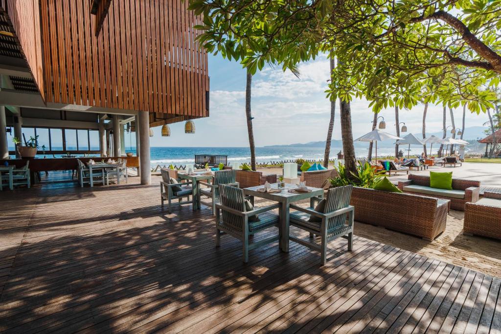 An outdoor seating area features wooden tables and chairs under a canopy of lush greenery. The ocean is visible in the background, with beach umbrellas and loungers placed comfortably on the sand, framed by palm trees and a clear sky.