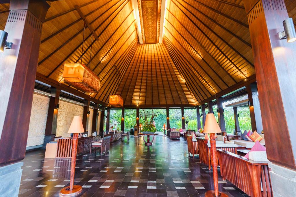 The open-air lobby features a high, thatched roof with warm lighting, enhancing the spacious ambiance. Natural materials like wooden beams and stone tiles are present on the floor. Lush greenery is visible outside through large windows, contributing to a serene atmosphere.