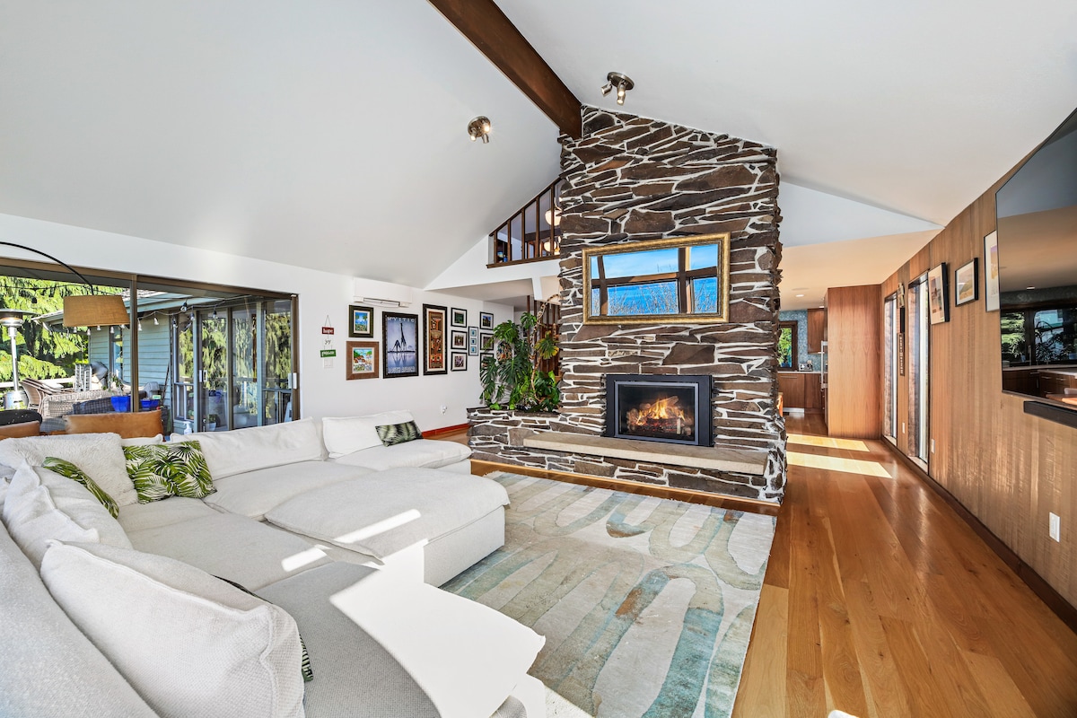 A spacious living room features a large sectional sofa arranged around a modern stone fireplace. Natural light floods the space through floor-to-ceiling windows, offering glimpses of the surrounding greenery. Colorful artwork adorns the walls, adding character to the inviting area.
