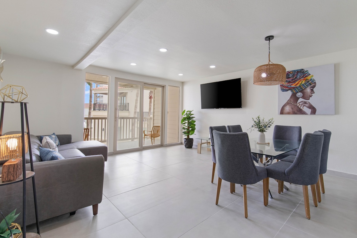 A modern living and dining area features a glass table surrounded by gray upholstered chairs. A cozy sectional sofa is positioned nearby, with a decorative lamp adding warmth. Natural light floods the space through sliding glass doors that lead to a balcony, complementing the neutral decor.