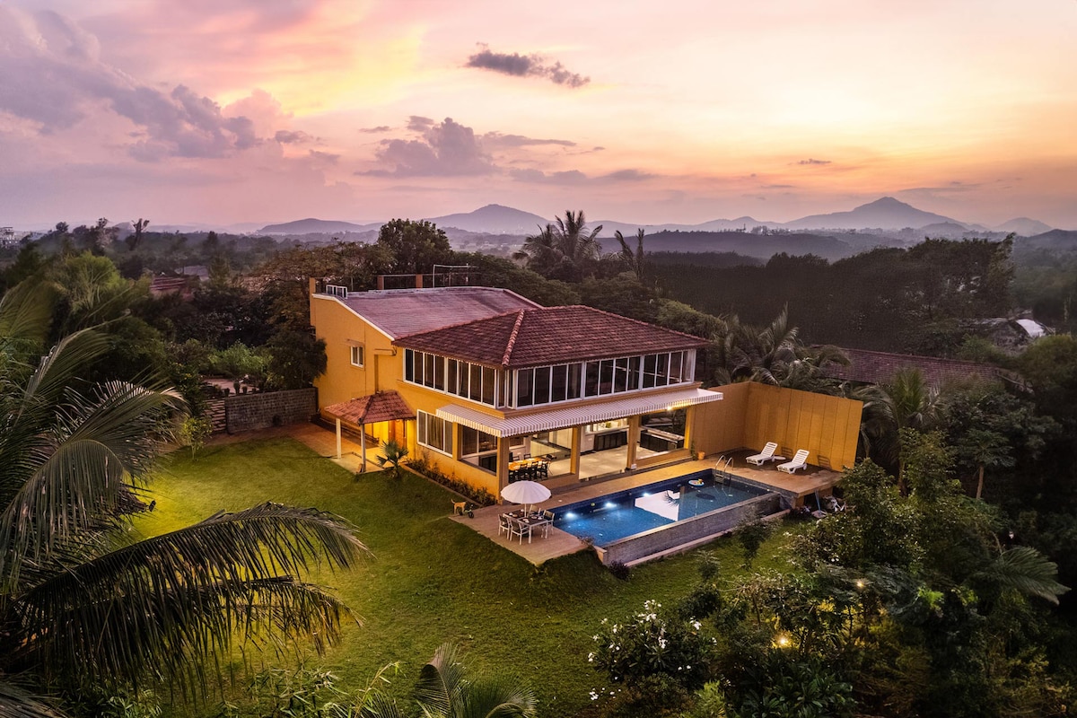 The villa's exterior showcases a spacious layout, with large windows reflecting the evening sky. A private pool and a well-maintained lawn can be seen outside, surrounded by lush greenery, while distant hills frame the background, contributing to a serene countryside setting.