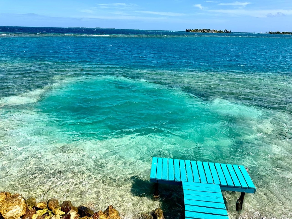 A wooden dock painted in turquoise extends over clear, shallow waters, providing direct access to the sea. Beneath the surface, varying shades of blue and green are visible, along with a rocky shoreline along the edge. Distant islands can be seen on the horizon.