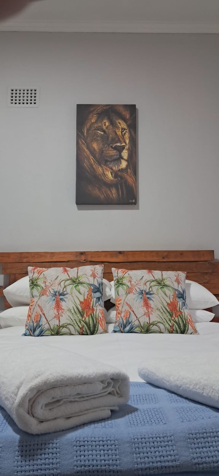 The sleeping area features a wooden headboard against a grey wall, adorned with decorative pillows showcasing vibrant floral designs. A piece of wall art hangs above the bed, complemented by neatly arranged white towels on a textured blanket.