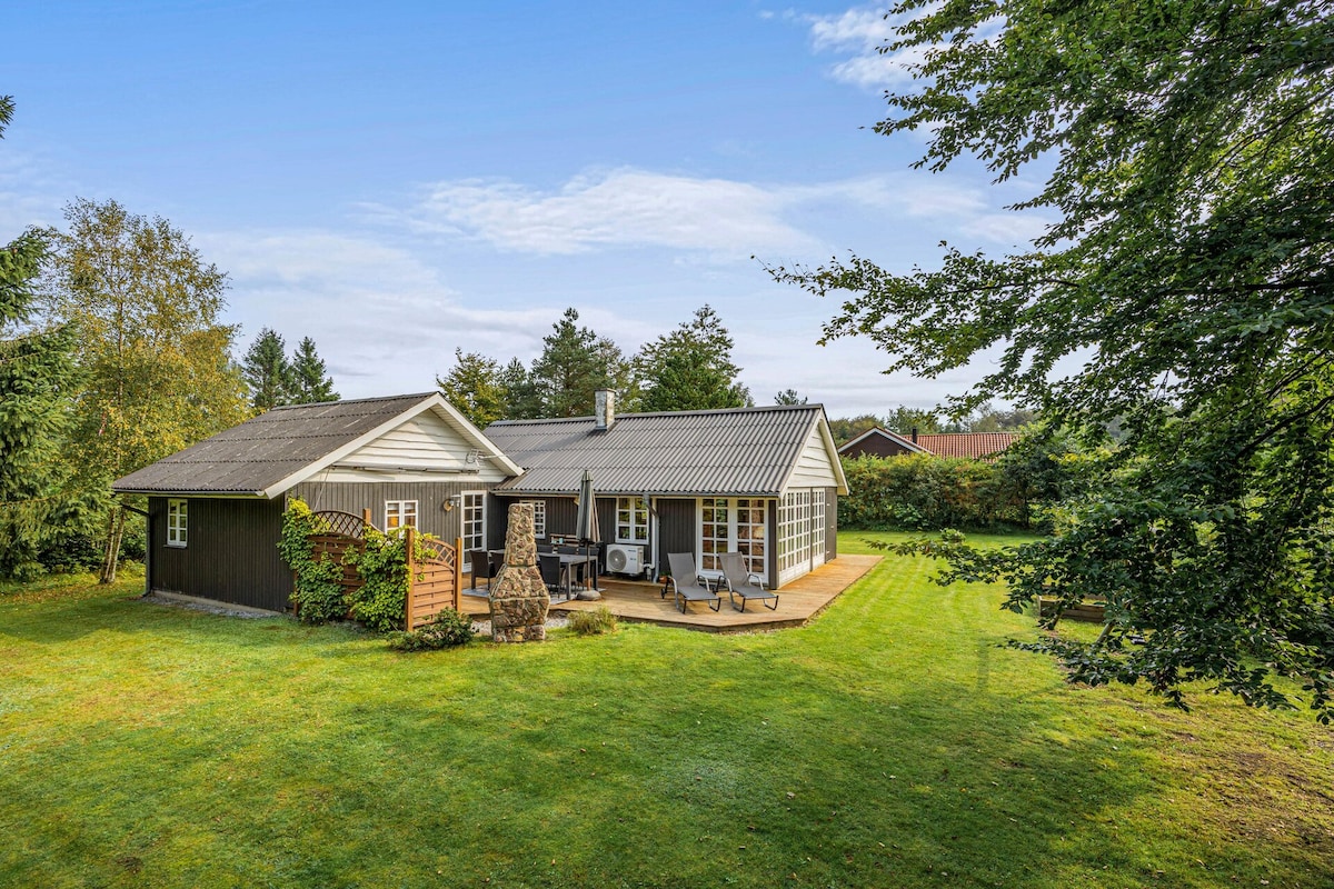 The exterior of the summerhouse is surrounded by lush greenery, with a spacious lawn. Outdoor seating is arranged on the terrace, featuring a grill and a cozy fireplace. The house's design combines wood and glass, blending with the natural surroundings.