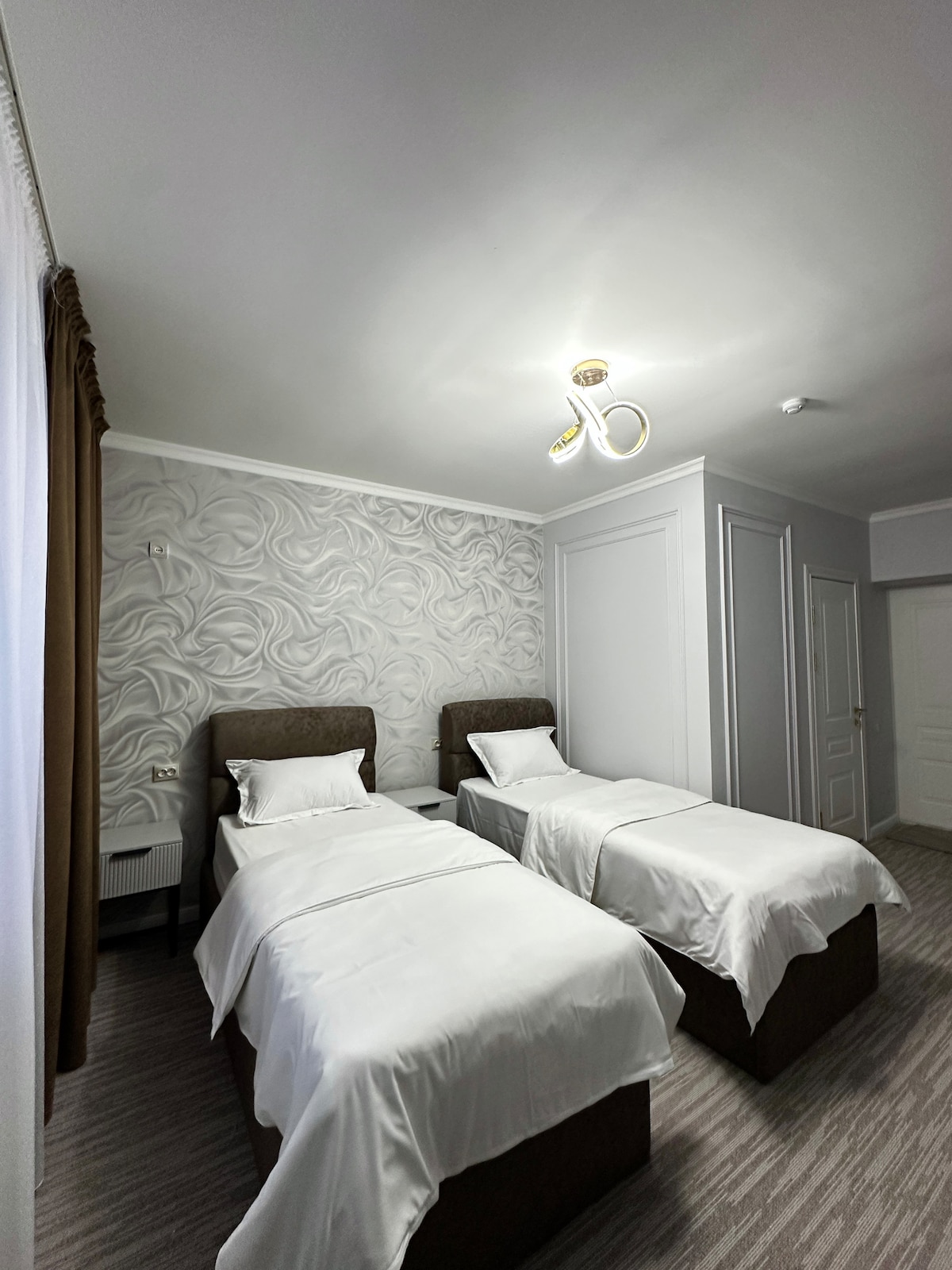 A well-appointed room features two separate beds with white linens, positioned with space between them. Textured light-colored wallpaper adorns one wall, while a soft chandelier illuminates the area. A small nightstand is placed beside each bed, contributing to the room's functional design.
