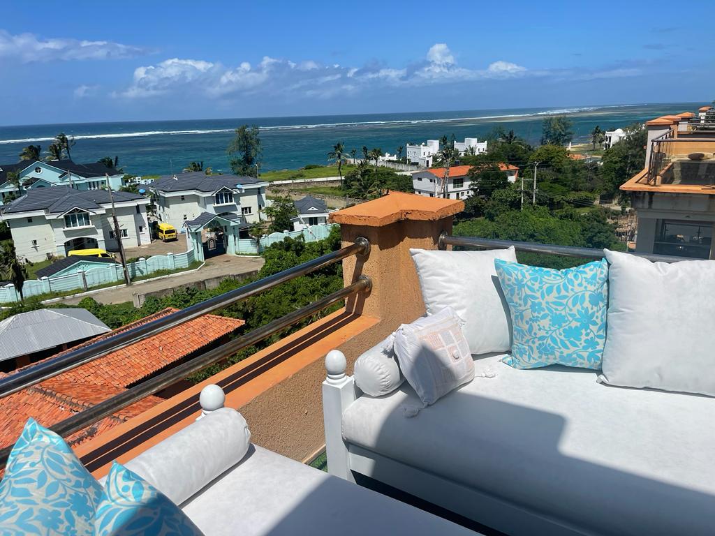 A spacious outdoor seating area is seen, featuring plush cushions in light blue and white. The background showcases a panoramic view of the ocean and surrounding coastal homes under a clear, sunny sky, enhancing the setting's tranquil environment.
