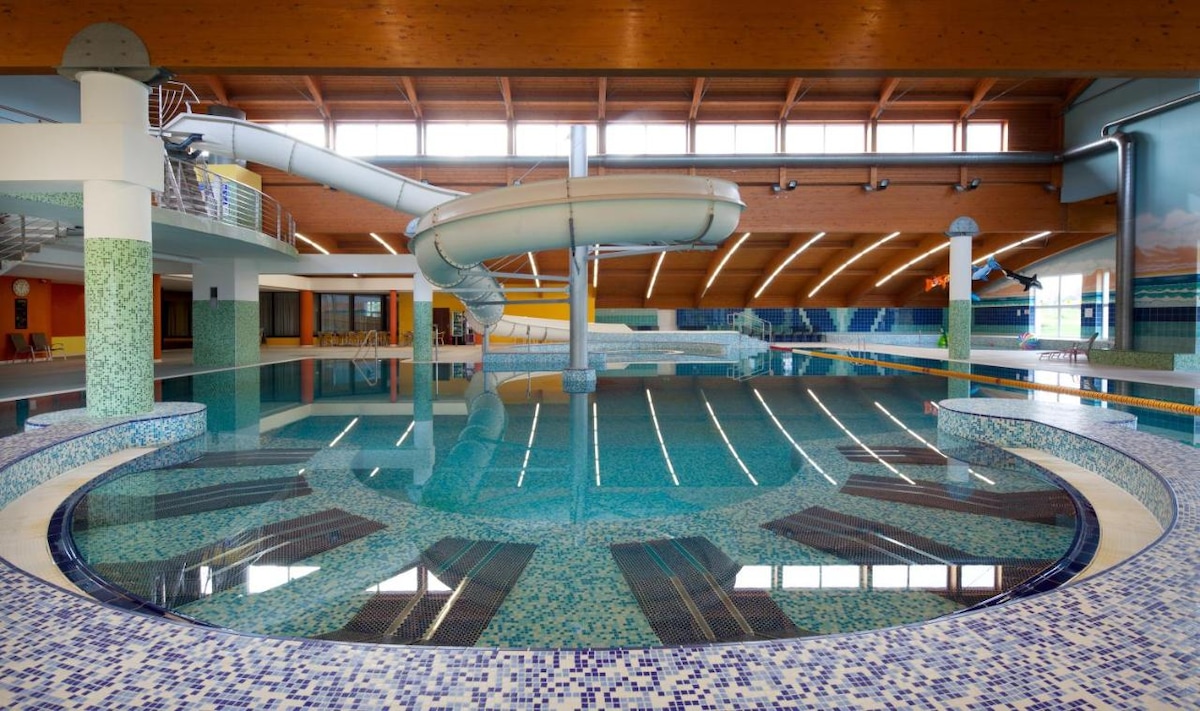A spacious indoor pool area is highlighted by a water slide, with colorful mosaic tiles decorating the edges. Natural light illuminates the clear water, and lounge chairs are neatly arranged along the poolside. The ceiling features wooden beams, adding to the inviting ambiance.