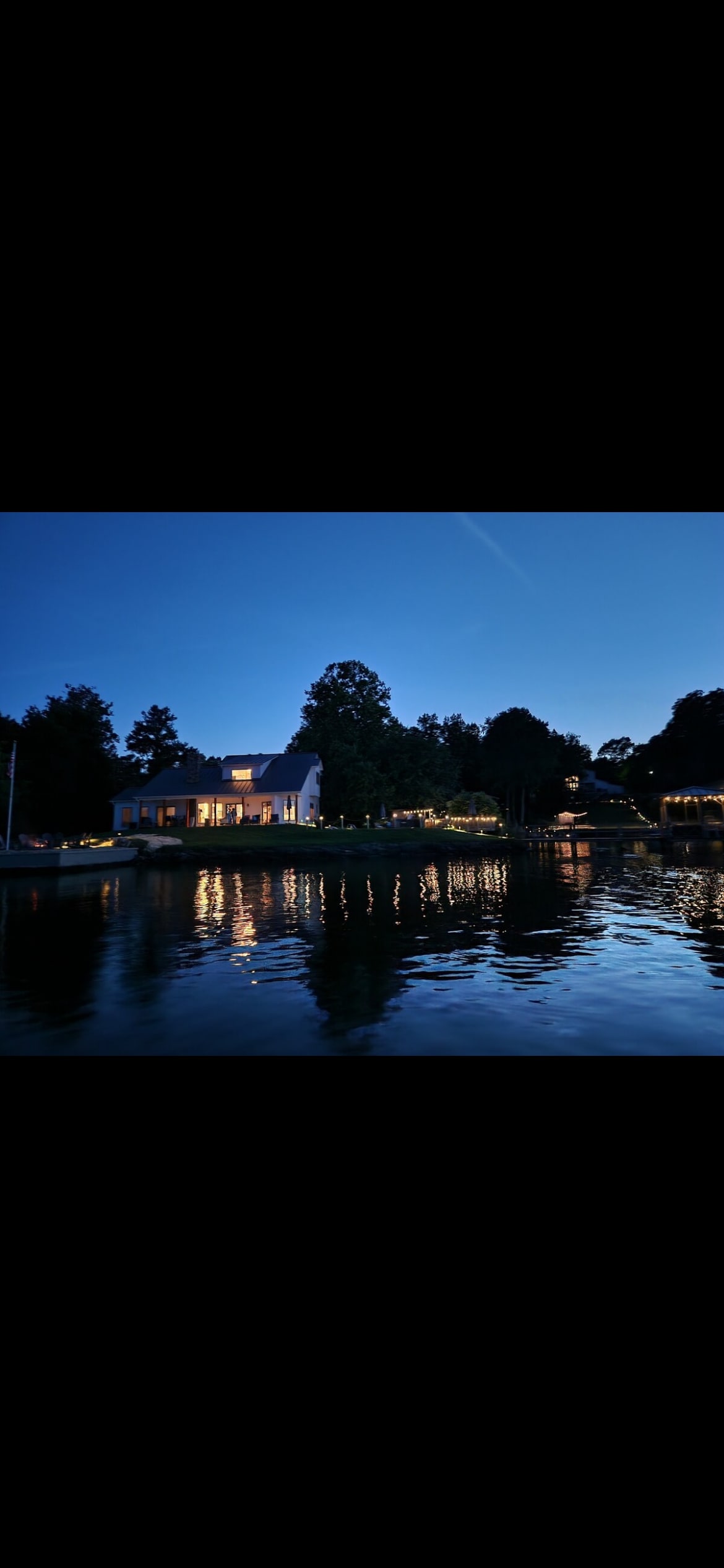 The exterior of the home is illuminated softly against the night sky, reflecting light on the serene water. Surrounding trees add a natural backdrop, enhancing the tranquil lakeside setting. The private boat dock is visible, providing direct access to the lake.