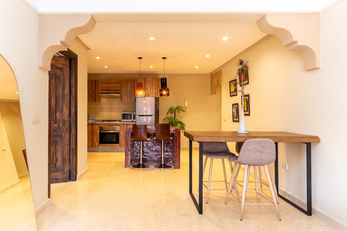 An open-concept kitchen and dining area features modern appliances and warm wood accents. A rectangular dining table is set for four, with stylish chairs positioned around it. Pendant lights hang above the kitchen island, adding a contemporary touch to the space.