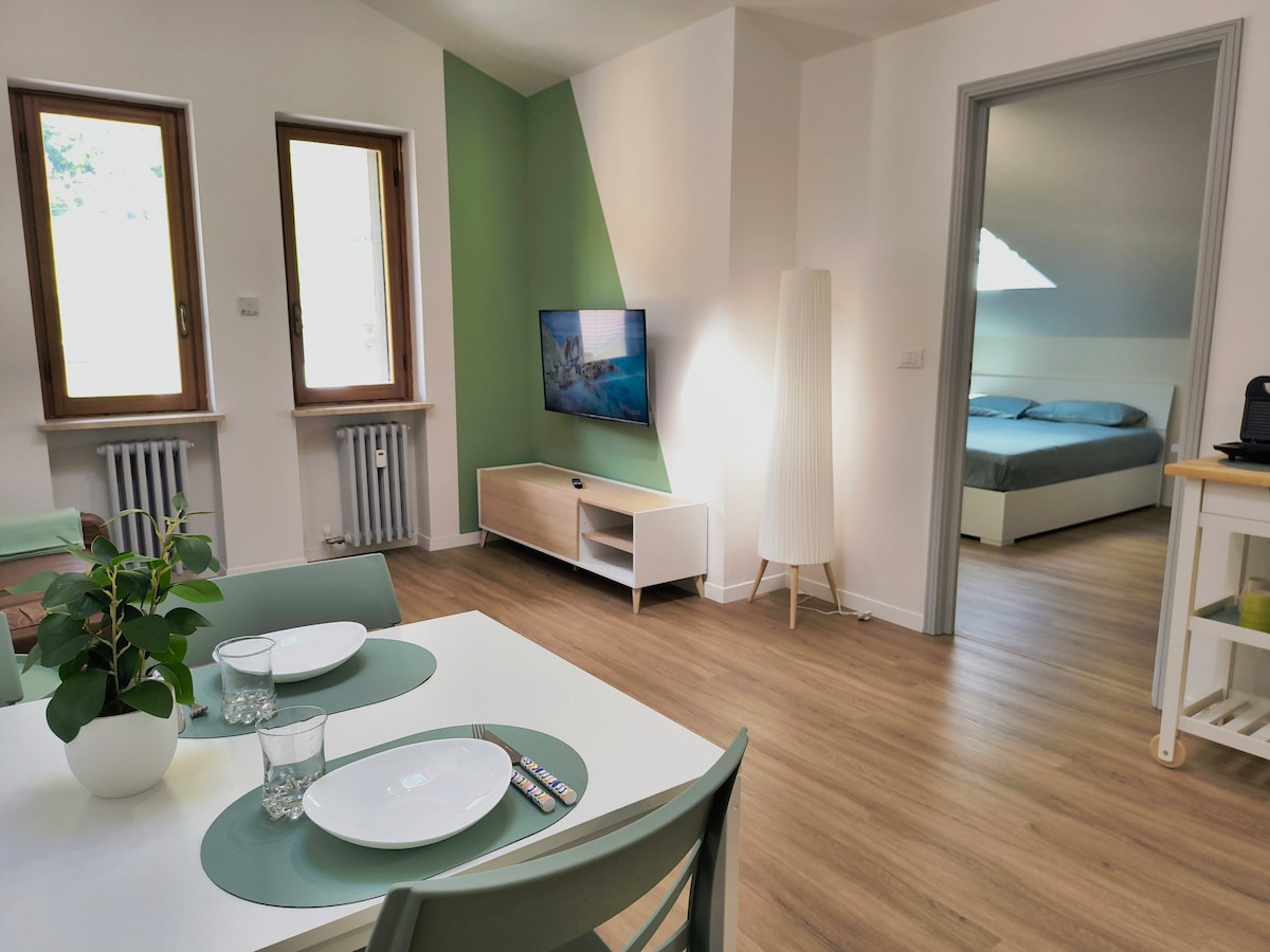 An open living area is presented, featuring light wood flooring and green accent walls. A dining table is set with plates and glasses, while a smart TV is mounted on the wall. Natural light floods through two windows, leading to the bedroom area with a double bed.