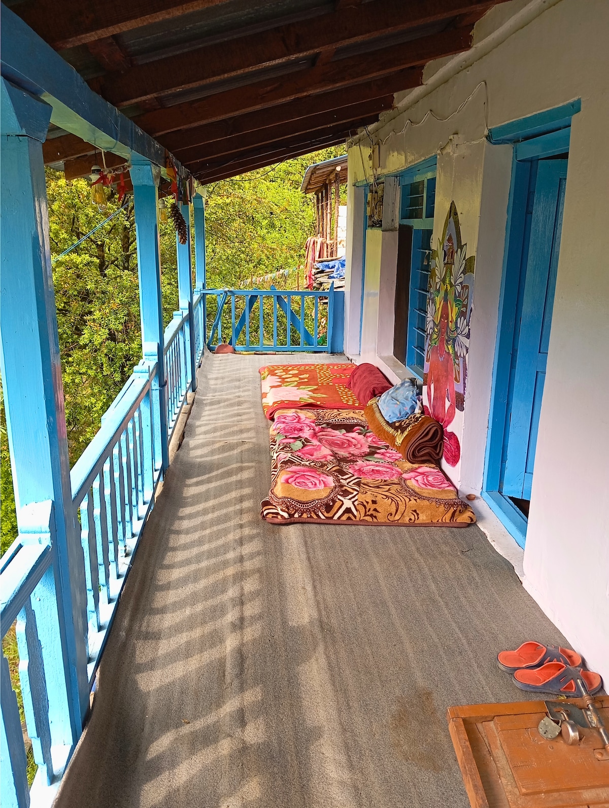 A covered balcony showcases comfortable seating arranged on a textured surface, complemented by colorful textiles. Bright blue wooden railings frame the space, while greenery and natural light create an inviting atmosphere. An entrance to a room is visible, enhancing the area’s accessibility.