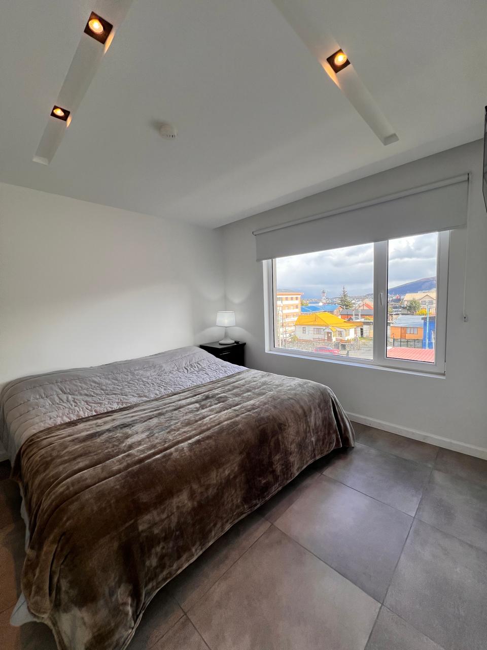 A modern bedroom is presented with a spacious bed covered in a textured brown blanket. A bedside table with a lamp is positioned next to the bed. Natural light enters through a large window, offering a view of the surrounding colorful buildings.