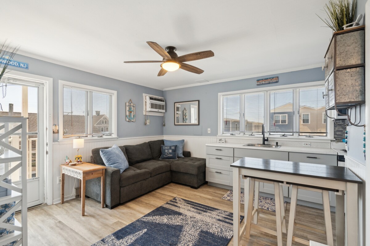 A light-filled living space features large windows offering views of the surroundings. A gray sectional sofa is positioned near the windows, complemented by a side table. A wooden dining table stands nearby, accompanied by decorative elements and a ceiling fan that enhances airflow.
