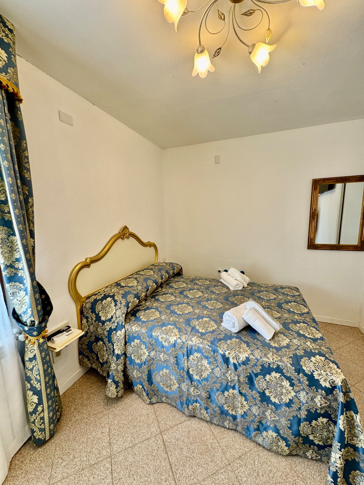 A cozy sleeping area features a large bed covered in a floral-patterned blue bedspread. Fresh towels are neatly arranged on the bed. Natural light enters through a window adorned with patterned curtains. A wall-mounted mirror reflects the room’s inviting layout.