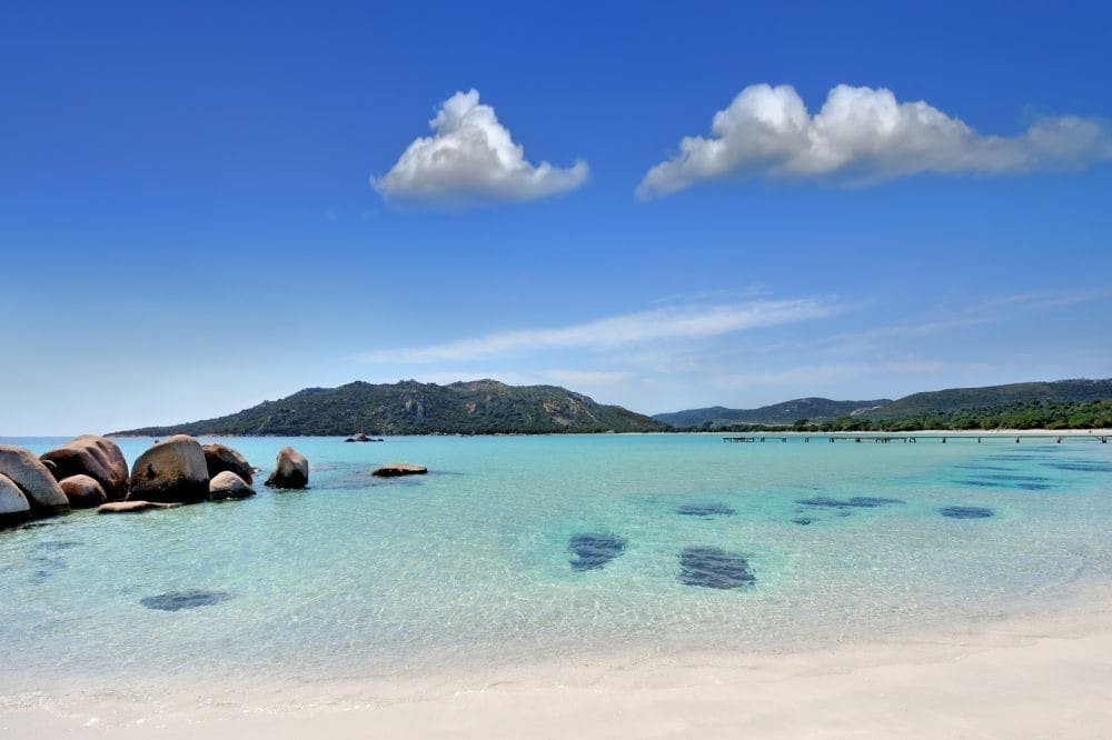 A serene beach scene is captured, showcasing gently lapping turquoise waters meeting a sandy shore. Large, smooth boulders are positioned along the water's edge, while a distant coastline features rolling hills under a clear blue sky with a few soft clouds.