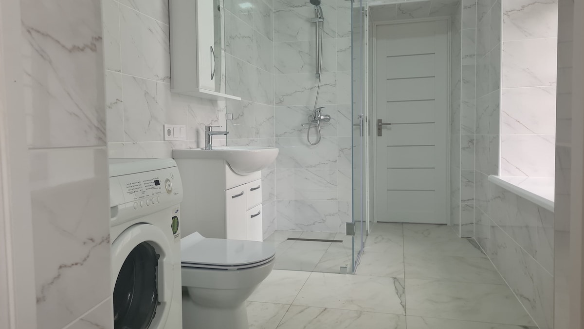 A modern bathroom is presented, featuring white marble tiles covering the walls and floor. A washing machine is placed beside a sleek basin with storage beneath. A glass shower enclosure stands on one side, while a window allows natural light to fill the space.