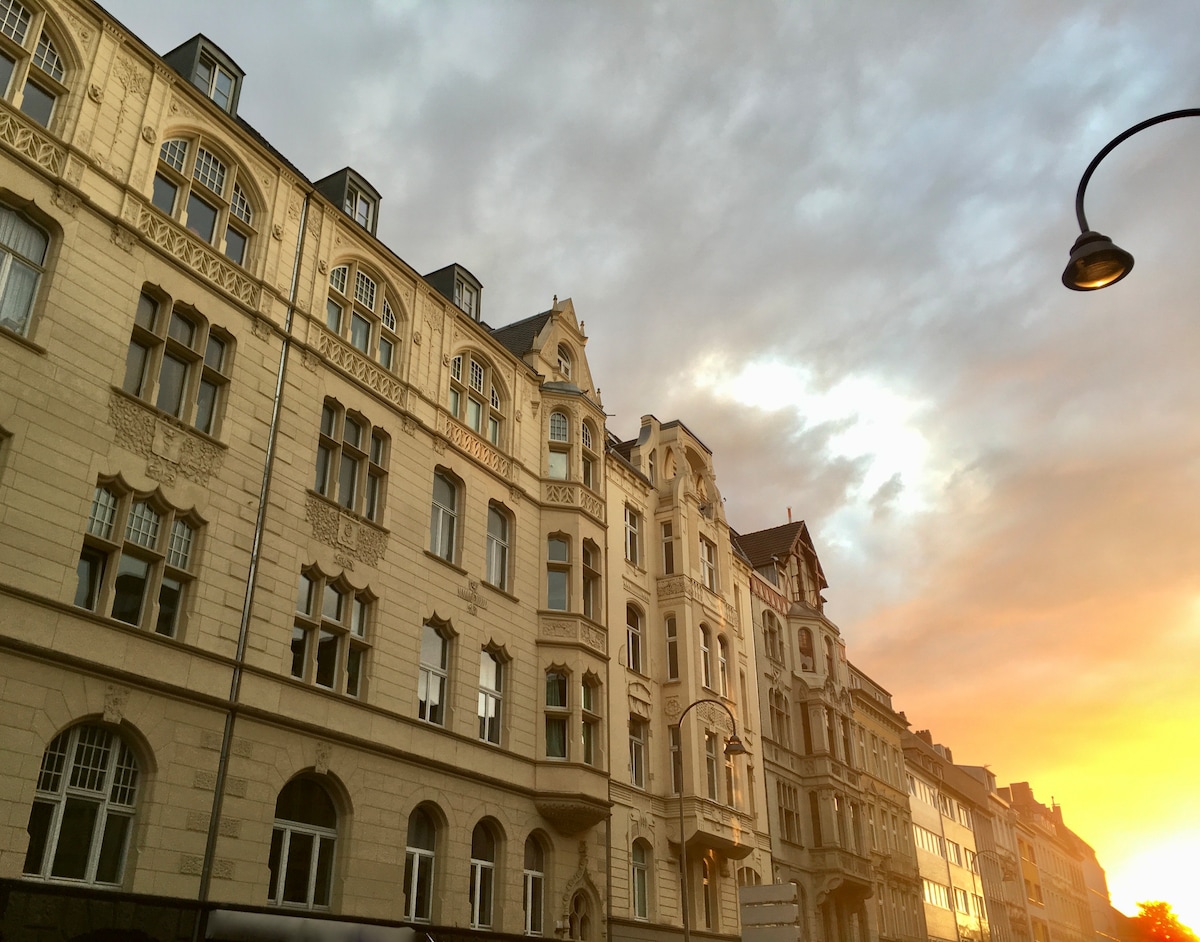 A historic building in the Jugendstil architectural style is shown, featuring decorative elements and large windows. The image captures a sunset, illuminating the façade with warm hues and accentuating the intricate details of the structure, set against a partially cloudy sky.