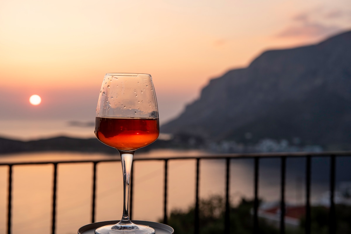 A glass of wine is placed on a table, showcasing droplets of condensation. The background features a sunset over a mountainous landscape, with warm orange and pink hues reflecting on the calm water. A wrought iron railing provides a border for the serene view.