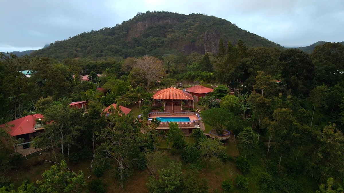Aerial view of a rustic cabin retreat nestled in lush greenery, surrounded by trees and hillside. The photo captures a central pool area with a red-tiled roof structure, complemented by various nearby buildings, all blending harmoniously with the natural landscape.