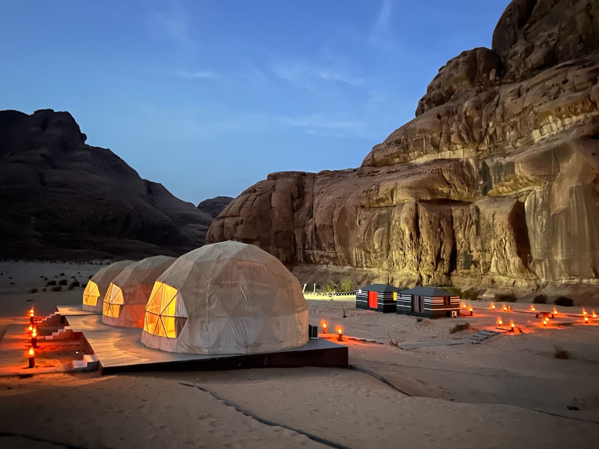 Three geodesic domes are positioned on a desert landscape, softly illuminated by warm lights. Surrounding the domes, the rugged rock formations rise steeply in the background, while gentle shadows are cast on the sandy ground, creating a serene evening ambiance.
