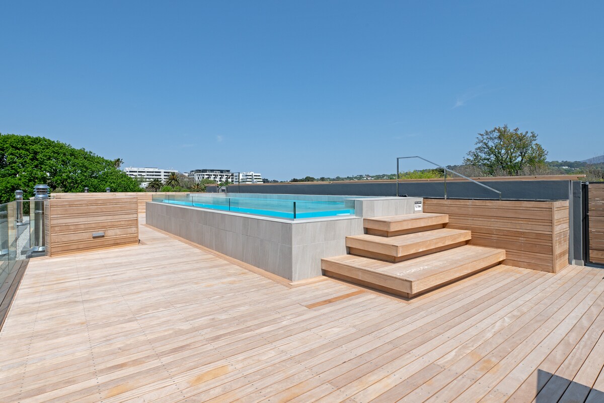 A rooftop pool area is displayed with a serene swimming pool surrounded by wooden decking. Steps lead down into the pool, complemented by glass railings. Lush greenery and a clear blue sky create a tranquil atmosphere.
