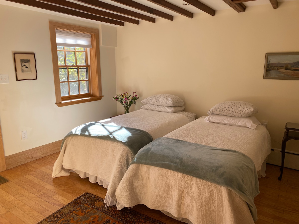 A room features two beds with soft, textured blankets in neutral tones. Natural light enters through a window with a wooden frame, illuminating the space. A small vase of flowers is placed on a nightstand beside the beds, contributing to a calm environment.