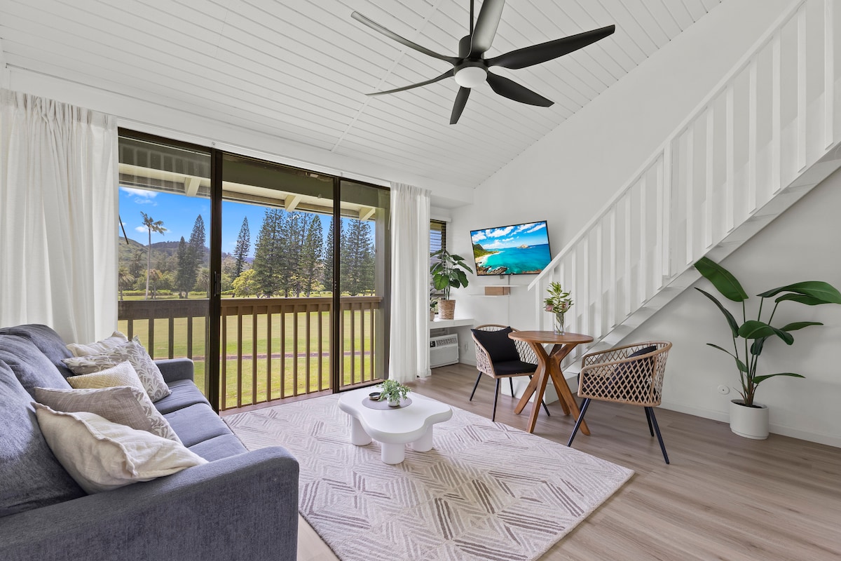 The living area features a comfortable sofa, a small coffee table, and a dining table for two. Large sliding doors provide ample natural light and showcase views of the golf course and mountains beyond. Ceiling fan and plants add to the ambiance.