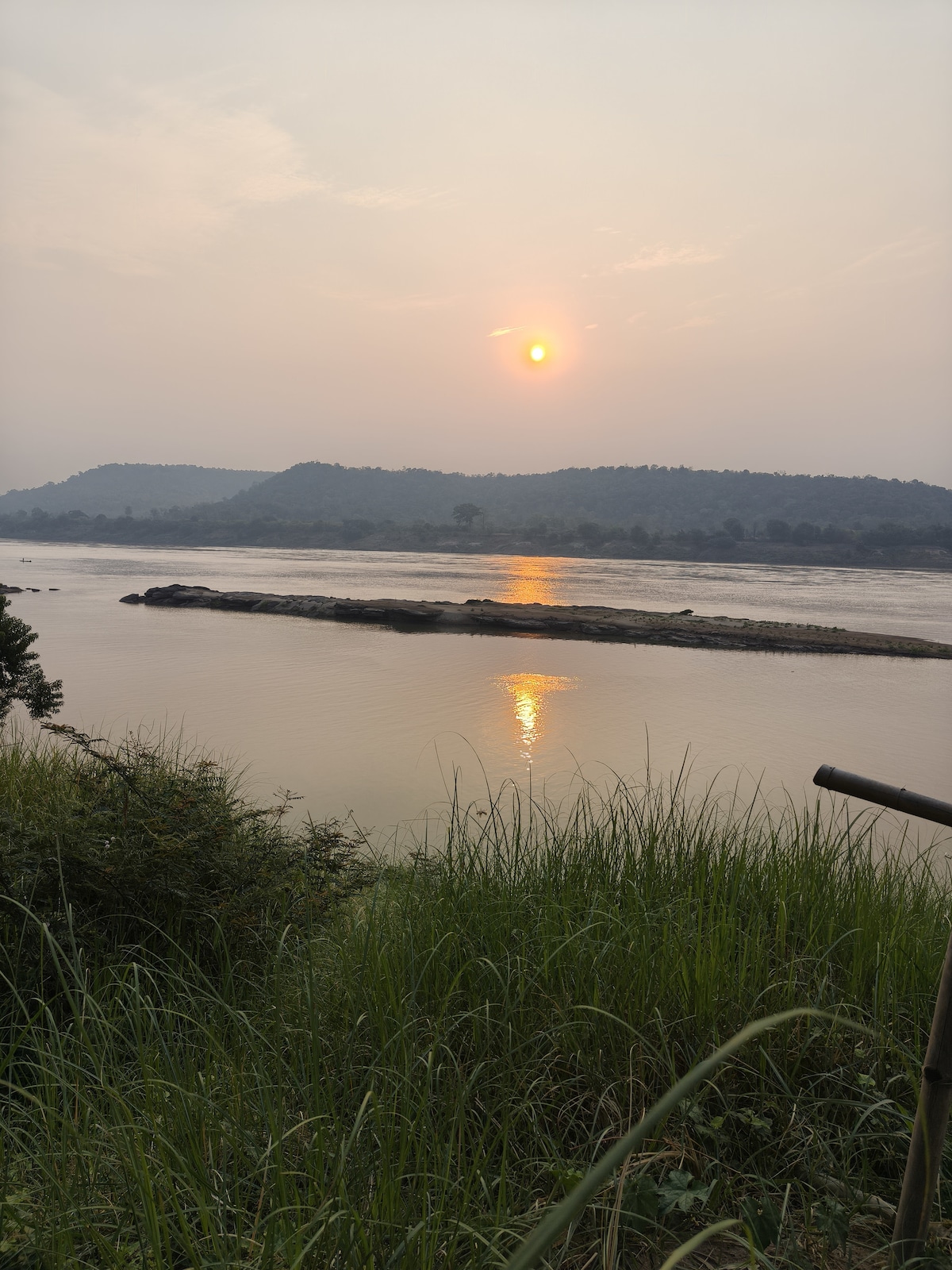 A serene view of the river during sunrise, with the sun rising above distant hills. The surface of the water reflects the soft orange glow, surrounded by lush green grass and gentle foliage. Small rock formations are visible in the river, enhancing the natural landscape.