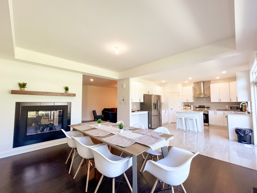 A spacious dining area is highlighted by a modern table surrounded by white chairs. A double-sided fireplace adds warmth, while the open kitchen showcases sleek cabinetry and stainless steel appliances, integrating cooking and dining spaces harmoniously.