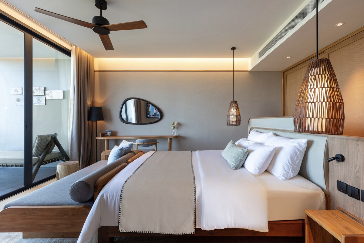 A light-filled bedroom features a spacious bed with crisp white linens and decorative pillows. Two woven pendant lights hang above warm wooden furnishings, while a floor-to-ceiling glass door opens to an outdoor seating area. A softly colored accent wall complements the soothing decor.