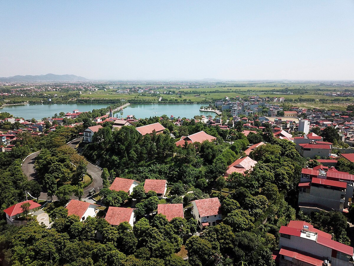 An aerial view of a vibrant community is presented, featuring neatly arranged houses with red roofs nestled among lush greenery. The surrounding landscape includes a peaceful body of water and distant mountainous terrain, creating a harmonious connection with nature.