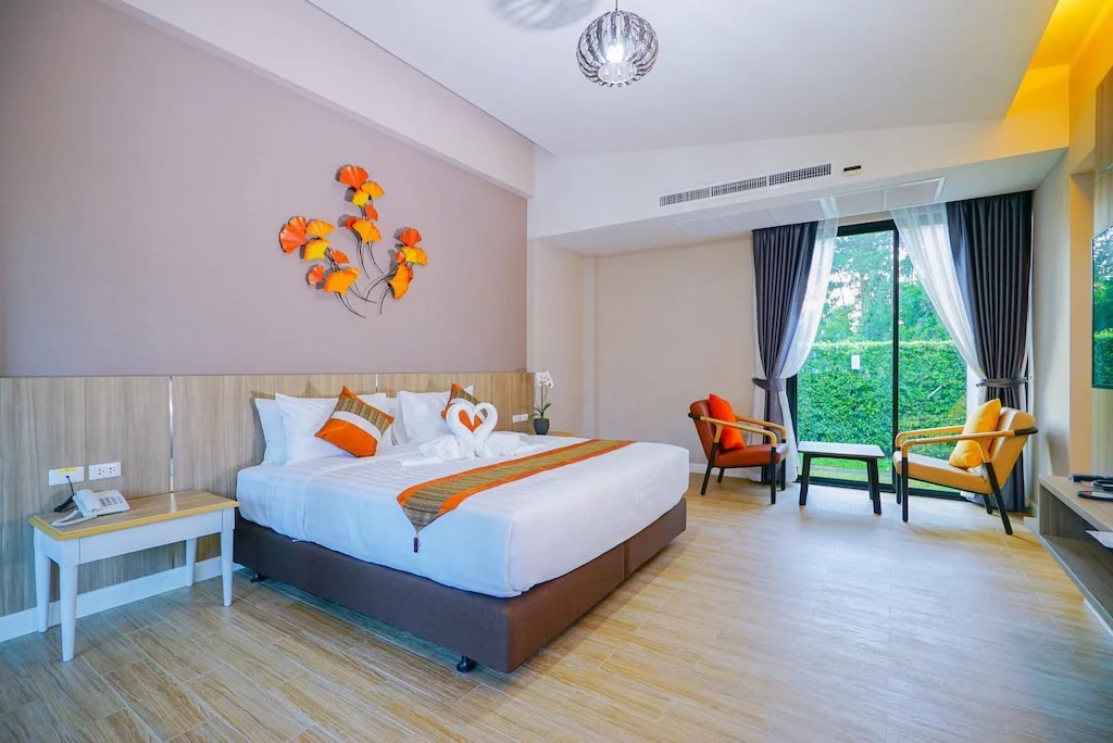 A spacious sleeping area features a large bed with decorative pillows and a towel art display. Accentuated by warm orange tones, the room includes seating with two chairs and a small table. Natural light shines through large windows, complemented by layered curtains.