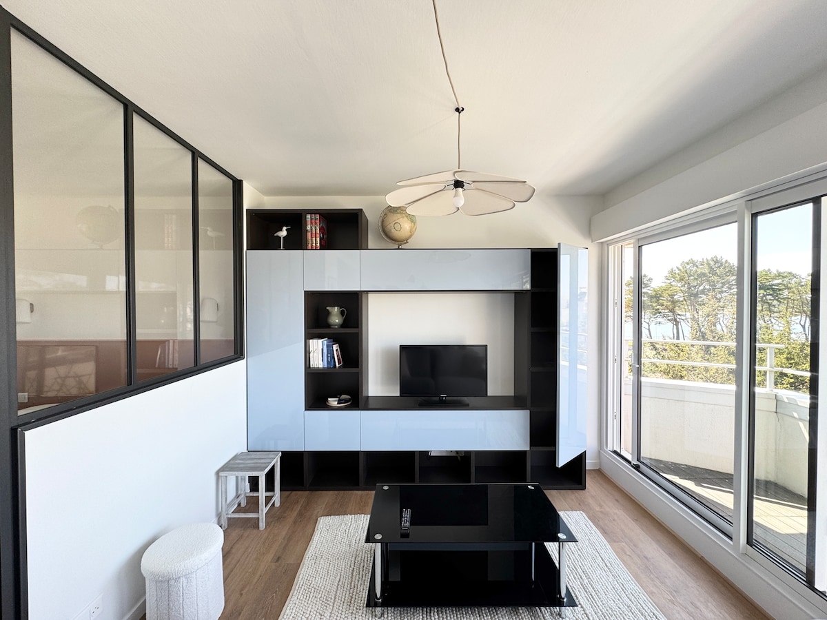 A bright living area features a modern black and white entertainment unit with shelving and a television. A dark coffee table is centered on a textured rug, complemented by a small white pouf. Large glass doors lead to a balcony with views of the ocean.