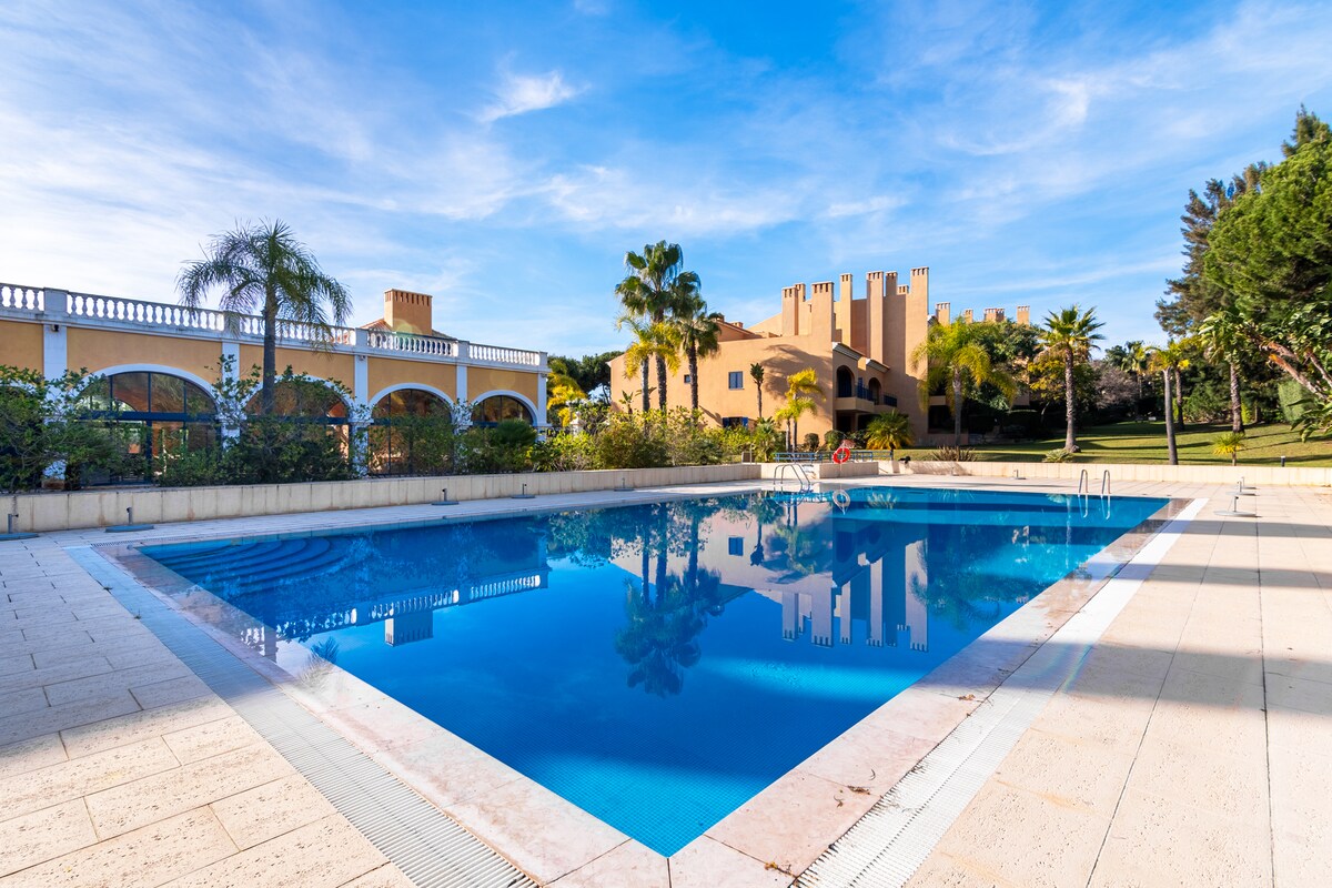 A large swimming pool is surrounded by a spacious patio and landscaped gardens. Sunlight reflects off the clear water, and several lounge chairs line the edge. The background features an elegant building with arched windows and palm trees, creating a serene and inviting atmosphere.
