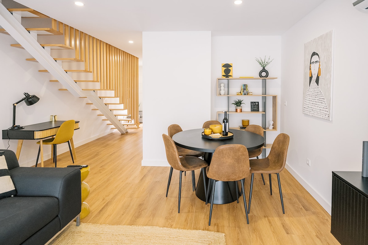 A modern dining area is presented with a round table surrounded by six brown leather chairs. A minimalist shelving unit displays decorative items. Light wood flooring complements the space, while a staircase with wooden slats leads to an upper level. A small desk and lamp are visible in the background.