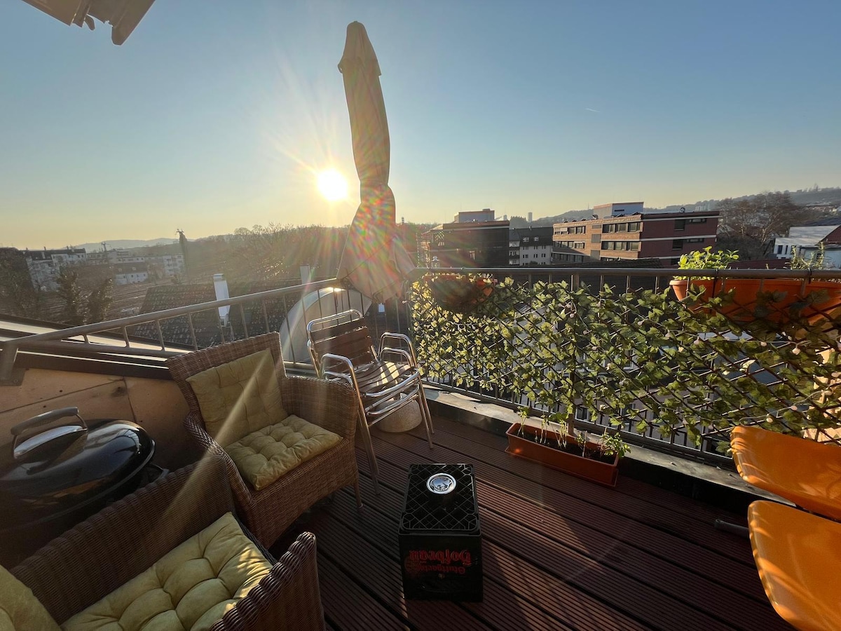 A rooftop terrace is bathed in golden sunlight, featuring comfortable seating arrangements with wicker chairs and a glass table. A portable grill is positioned nearby, and potted plants line the edge, creating an inviting outdoor space with panoramic views of the surrounding area.