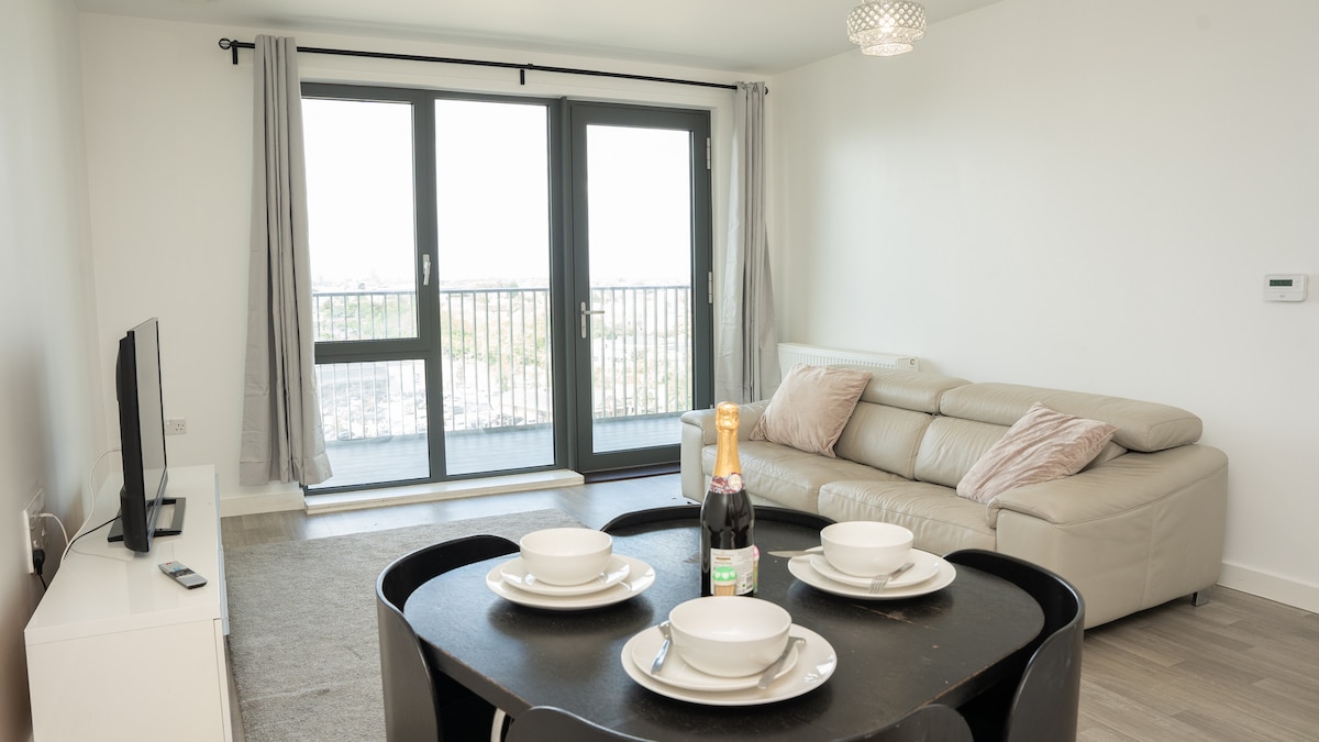 A modern living area features a light-colored sofa and a dark table set with dinnerware. A flat-screen TV is positioned against a white wall. Large glass doors lead to a balcony, framed by long, light curtains. A crystal chandelier adds a touch of elegance.