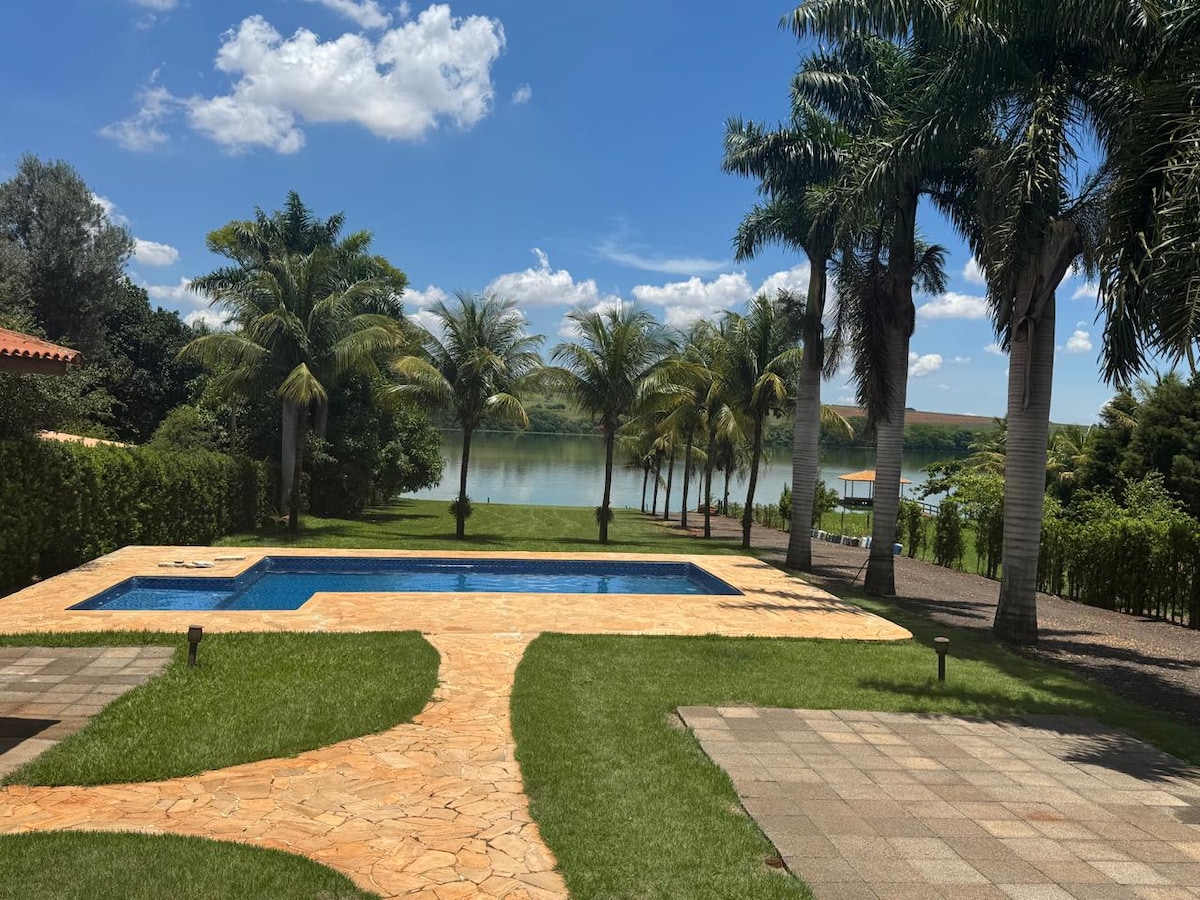 A serene outdoor area features a swimming pool surrounded by lush greenery and palm trees. The pathway leads towards the tranquil setting by the water, where reflection of the sky can be seen. A clear blue sky enhances the natural ambiance of the space.