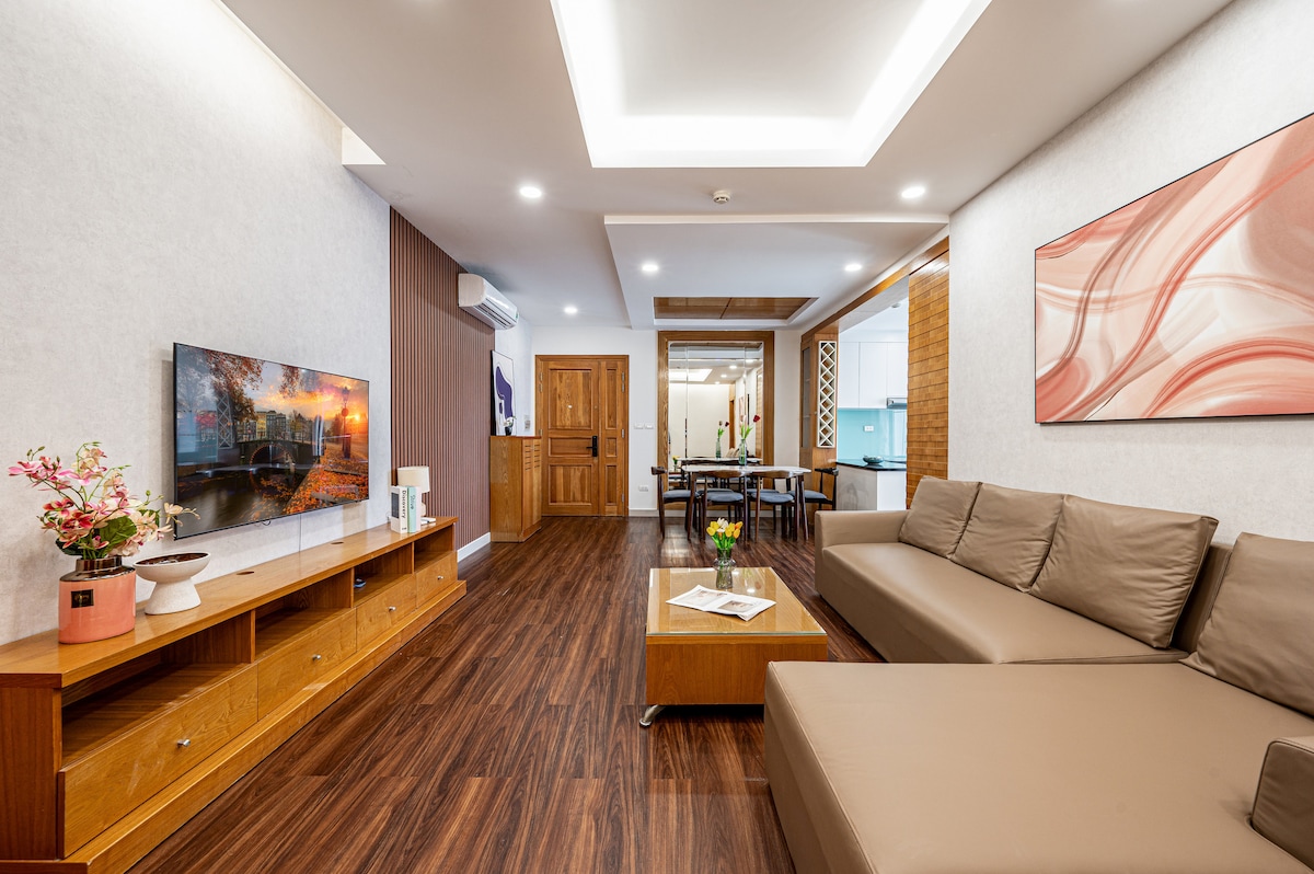 The living room features a modern sofa and a coffee table, with decorative items placed on a media console below a wall-mounted LED TV. Light-colored walls and a warm wood floor enhance the spaciousness, while natural light fills the area from the doorway leading to the dining space.