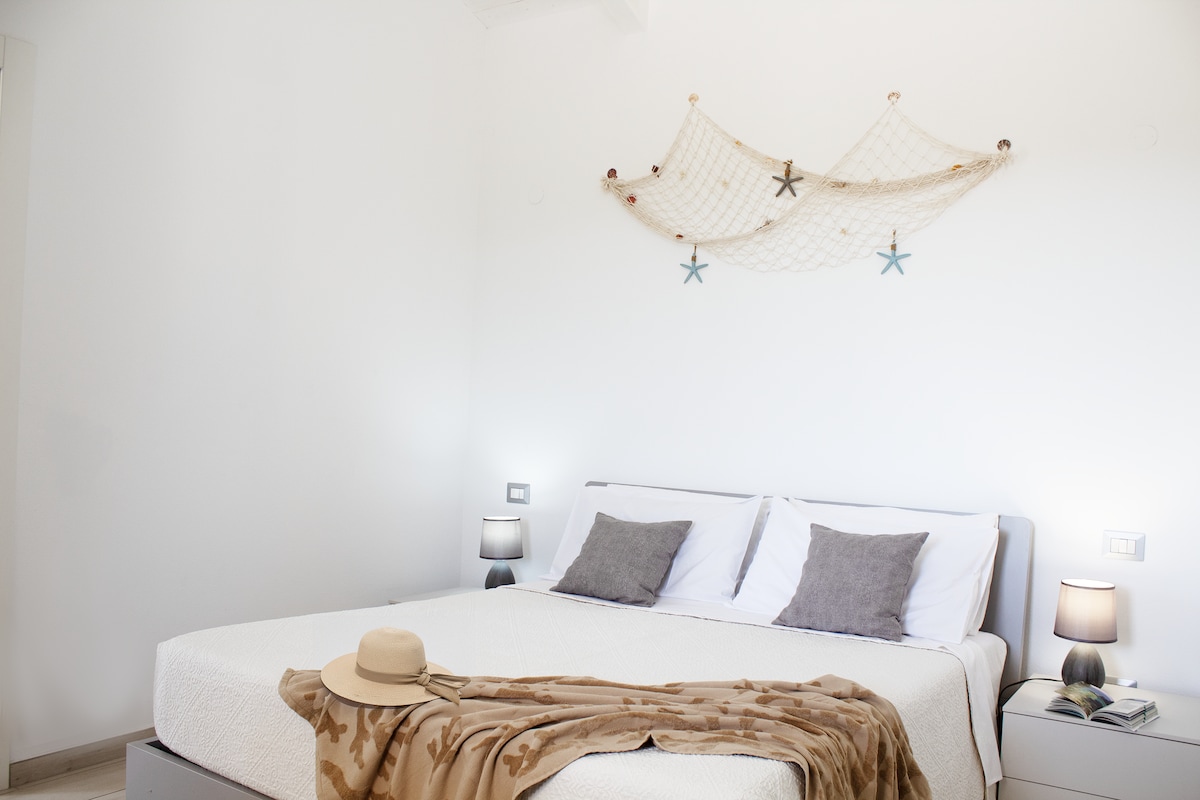 A bright and spacious bedroom is depicted, featuring a large bed with white linens and decorative pillows. Soft bedside lamps provide gentle lighting. A decorative net with starfish is displayed above the bed, adding a coastal touch to the airy atmosphere.