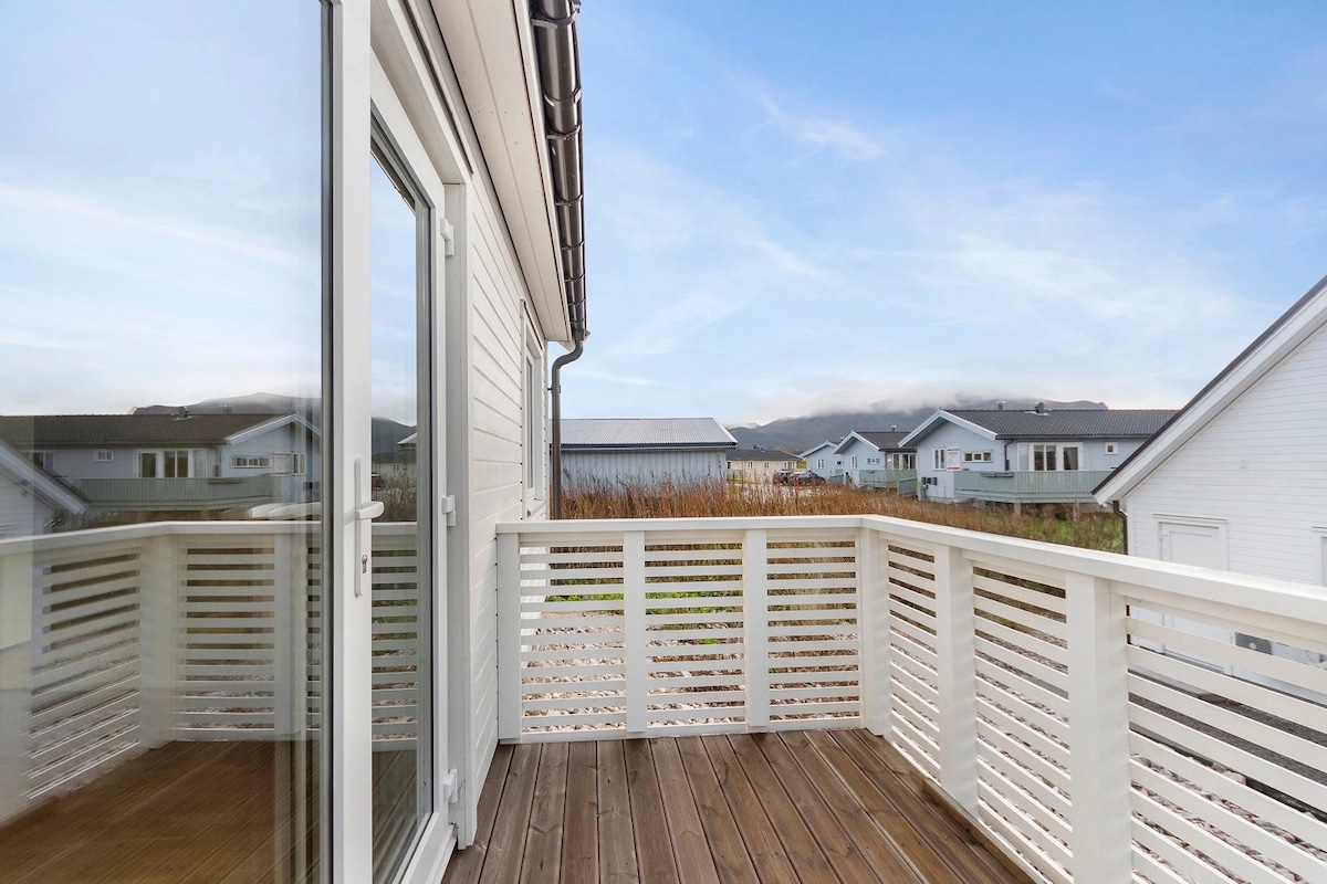 A spacious balcony is displayed, featuring wooden flooring and a white railing. Scenic views of neighboring houses and distant hills are visible under a clear blue sky, providing a tranquil outdoor space for relaxation.