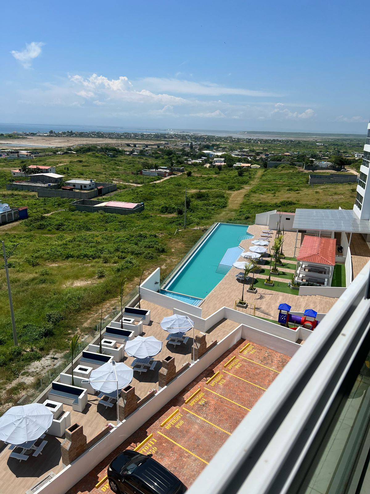 An aerial view captures a swimming pool surrounded by lounge chairs and umbrellas, showcasing an inviting outdoor space. Lush greenery and distant urban sights provide a contrasting backdrop, while the sun illuminates the area, highlighting the poolside layout and seating arrangements.