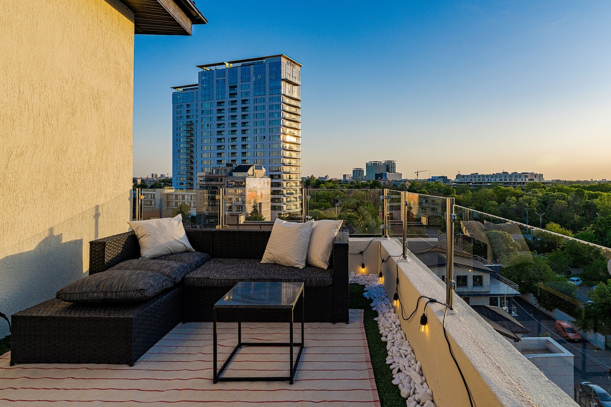 A terrace features a dark wicker sofa adorned with light-colored cushions, accompanied by a glass-topped table. The skyline showcases modern buildings in the background, while a soft glow is provided by string lights along the railing. The space is perfect for relaxation and outdoor dining.