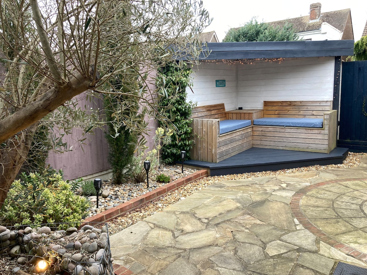 A private courtyard garden features a covered seating area with built-in wooden benches and blue cushions. The space is surrounded by greenery and decorative stones, with soft lighting illuminating the path. Mature trees blend with pebbled landscape elements, creating a serene outdoor retreat.