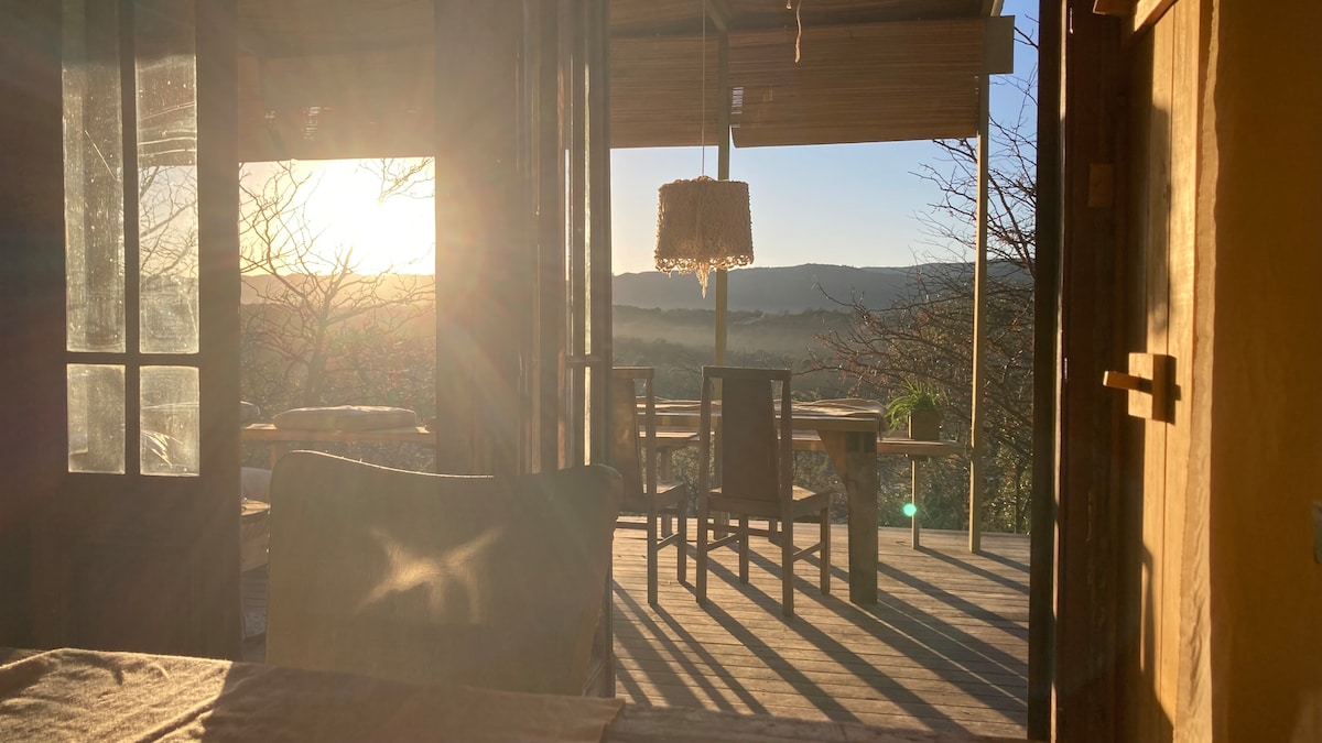 Morning light filters through open doors, illuminating the wooden deck and providing expansive views of the surrounding hills. A rustic chandelier hangs above a table set for outdoor dining, enhancing the connection with nature. Subtle shadows are cast across the floor, creating a peaceful ambiance.