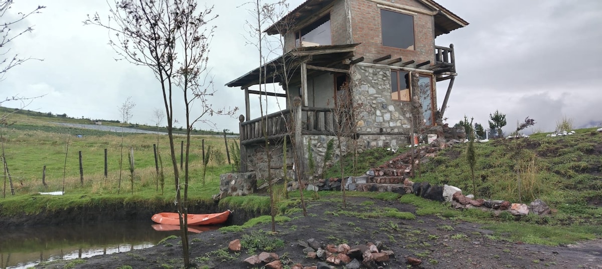 A rustic two-story building is surrounded by green grass and trees. A small body of water is visible nearby, with an orange kayak positioned near the shore. Steps made of stones lead up to the entrance, framed by a wooden balcony that offers an outdoor seating area.