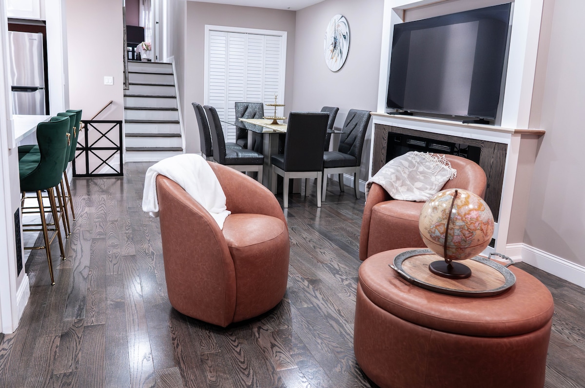 A modern living area is presented with warm wooden flooring. Two brown accent chairs are positioned beside a circular ottoman featuring a globe on it. A dining space with dark chairs and a staircase in the background adds depth to the open layout, while a large television is mounted on the wall.